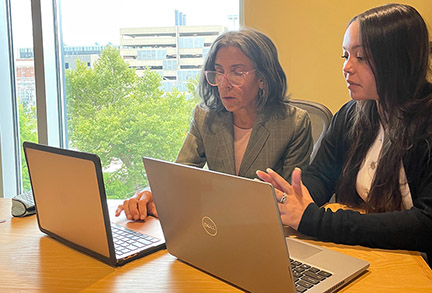 women sitting at table, looking at their laptops, while one explains something to the other